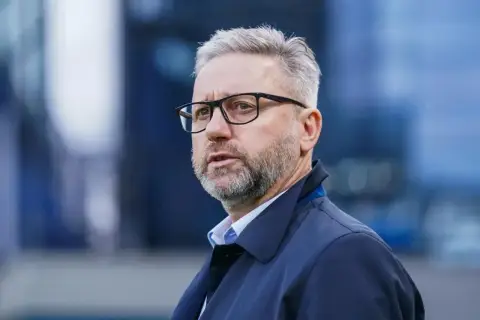 Brzęczek trener w okularach, z siwą brodą i włosami, w granatowej kurtce, patrzy w bok.