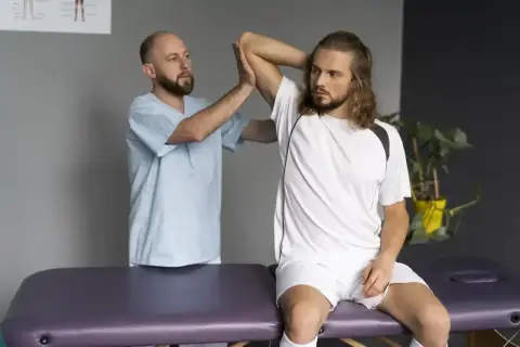 Fizjoterapeuta bada naciągnięty bark młodego sportowca siedzącego na kozetce.