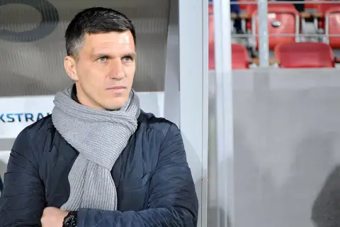 Robert Podoliński, trener w szaliku, spogląda w bok na tle stadionu z czerwonymi siedzeniami.